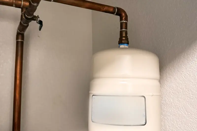 Small white expansion tank hanging from a copper water line, a second copper line and valve running beside it