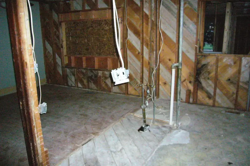 A gutted room stripped back to old wall boards and bare subfloor, with wiring hanging loose and water supply and drain pipes sticking up from the floor