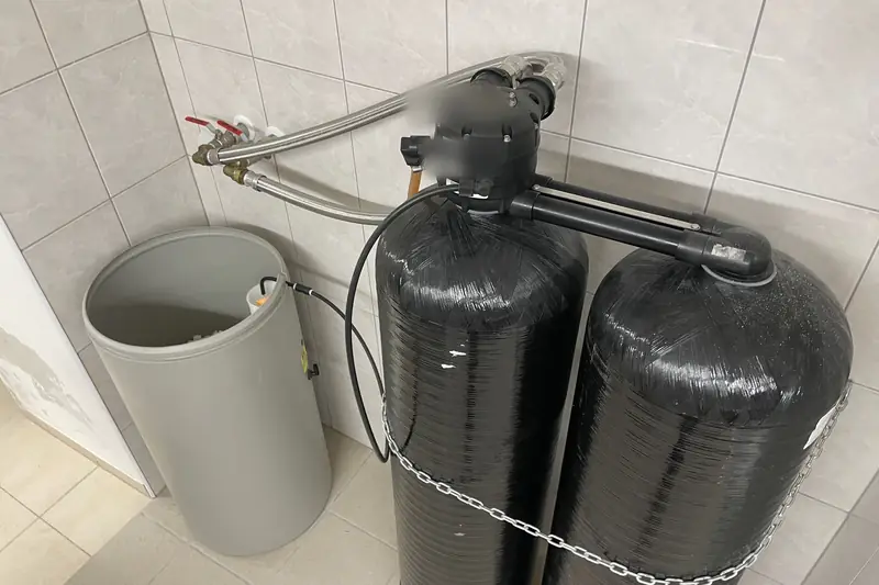 Two black water softener tanks with a control valve on top and a grey brine tank beside them, joined by braided hoses to the house water lines
